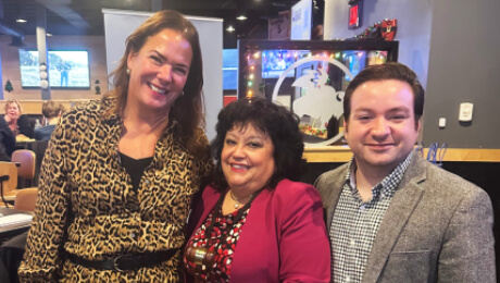Three individuals posing together in a lively restaurant setting, with festive decorations in the background.
